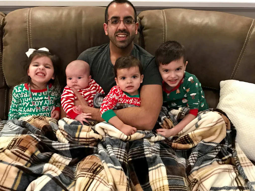 A smiling man sits on a couch with four young children in his lap and at his sides. The children wear red and green pajamas and a plaid blanket is over their laps. 