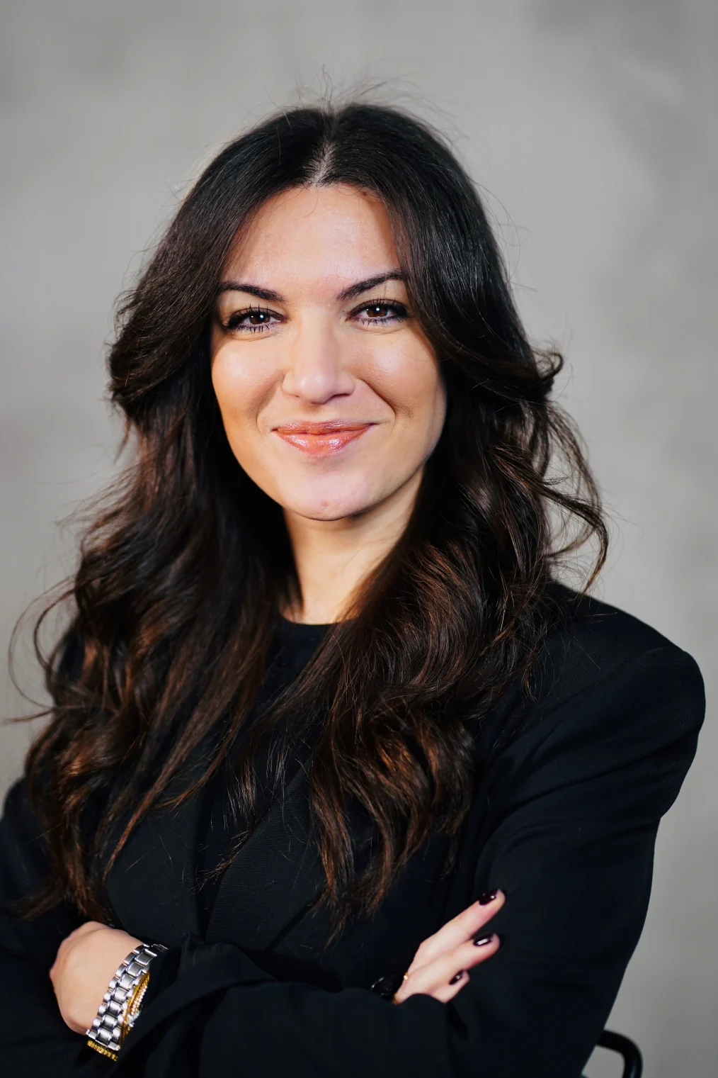 Ornella Cosomati, a woman with a light skin tone and long black hair, smiles at the camera. She has a closed lip smile and is wearing a black blazer. Her arms are crossed. 