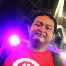 A medium skin toned Brown man with short dark hair is smiling slightly with his mouth closed. He is wearing a red t shirt with backpack straps showing on either side. There is a bright pink light shining behind him. 