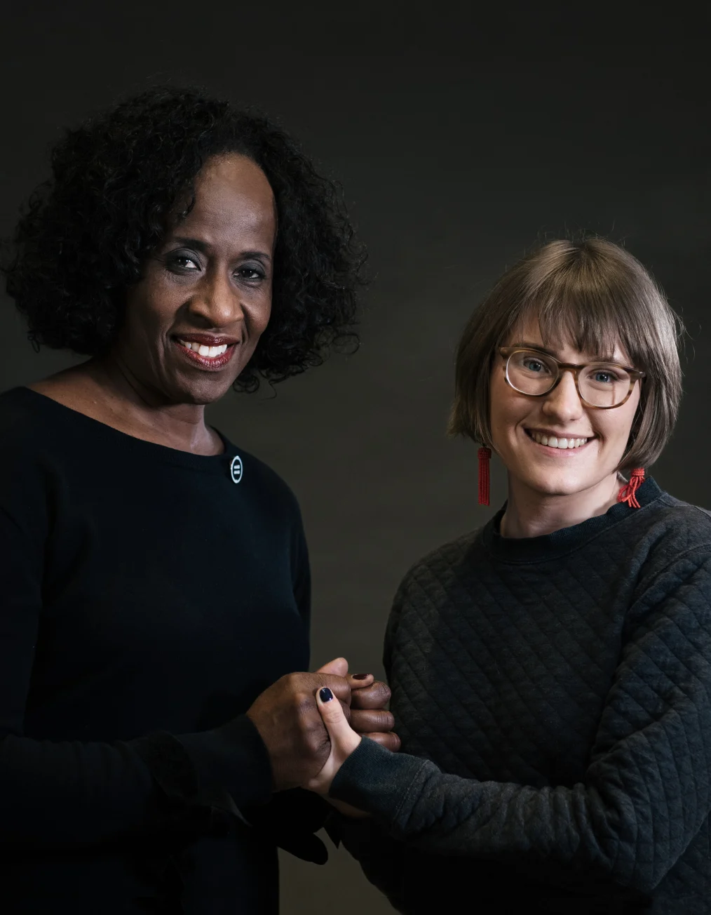 This picture shows two women with different skin tones grasping each other's hands and
smiling at the camera. One lady's skin tone is deep, while the other's is light.
