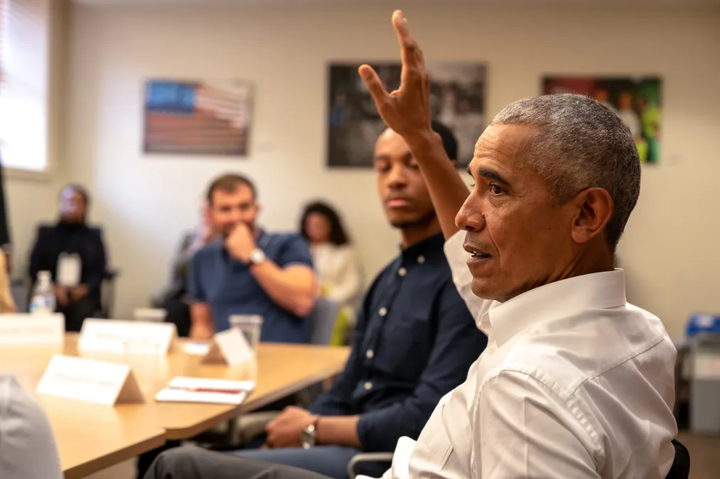 President Obama raises his hand to speak at a table full of people with various skin tones.
