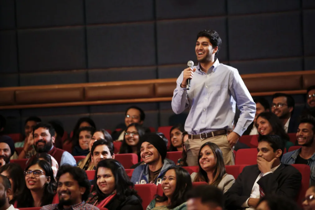 A man with a medium, warm golden skin tone with slicked back hair, a light purple button up shirt, and khakis, standing and speaking on a microphone in the middle of a seated audience.