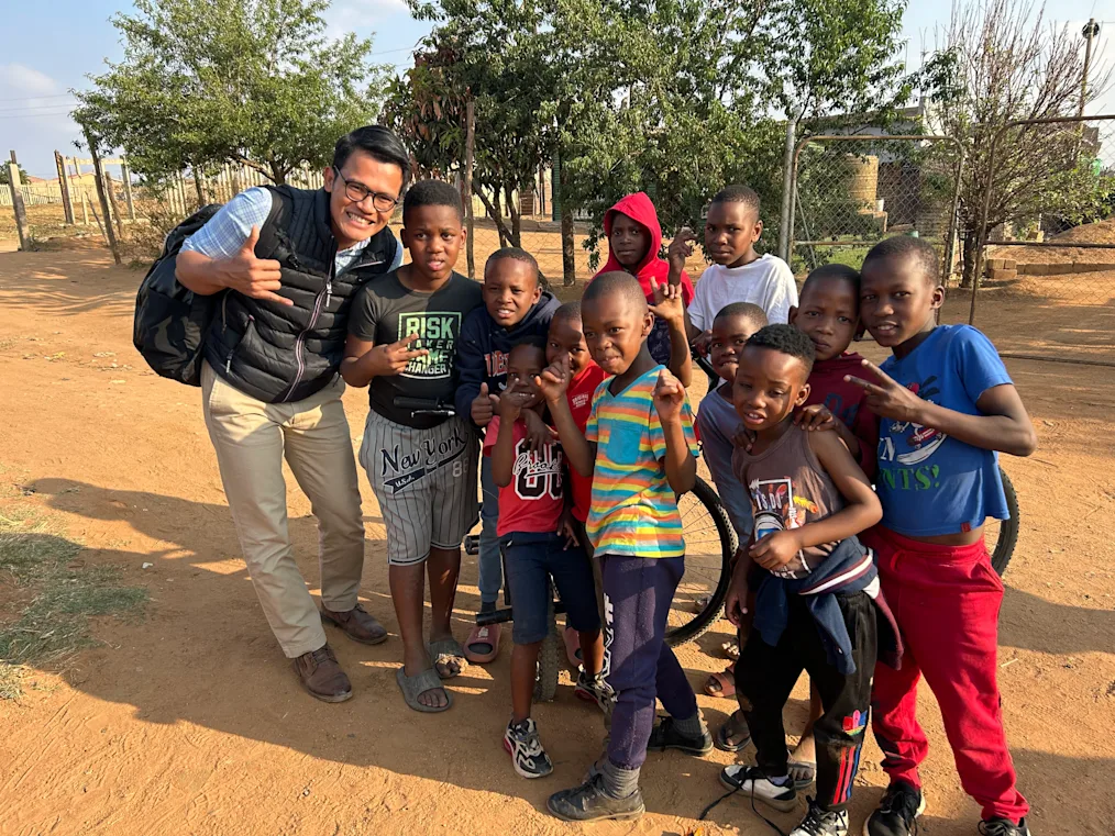 Charles Klaikaew, an Asian man with a light skin tone, poses next to a group of Black children with dark skin tones, during a trip to eMpumelelweni Township. All are smiling.