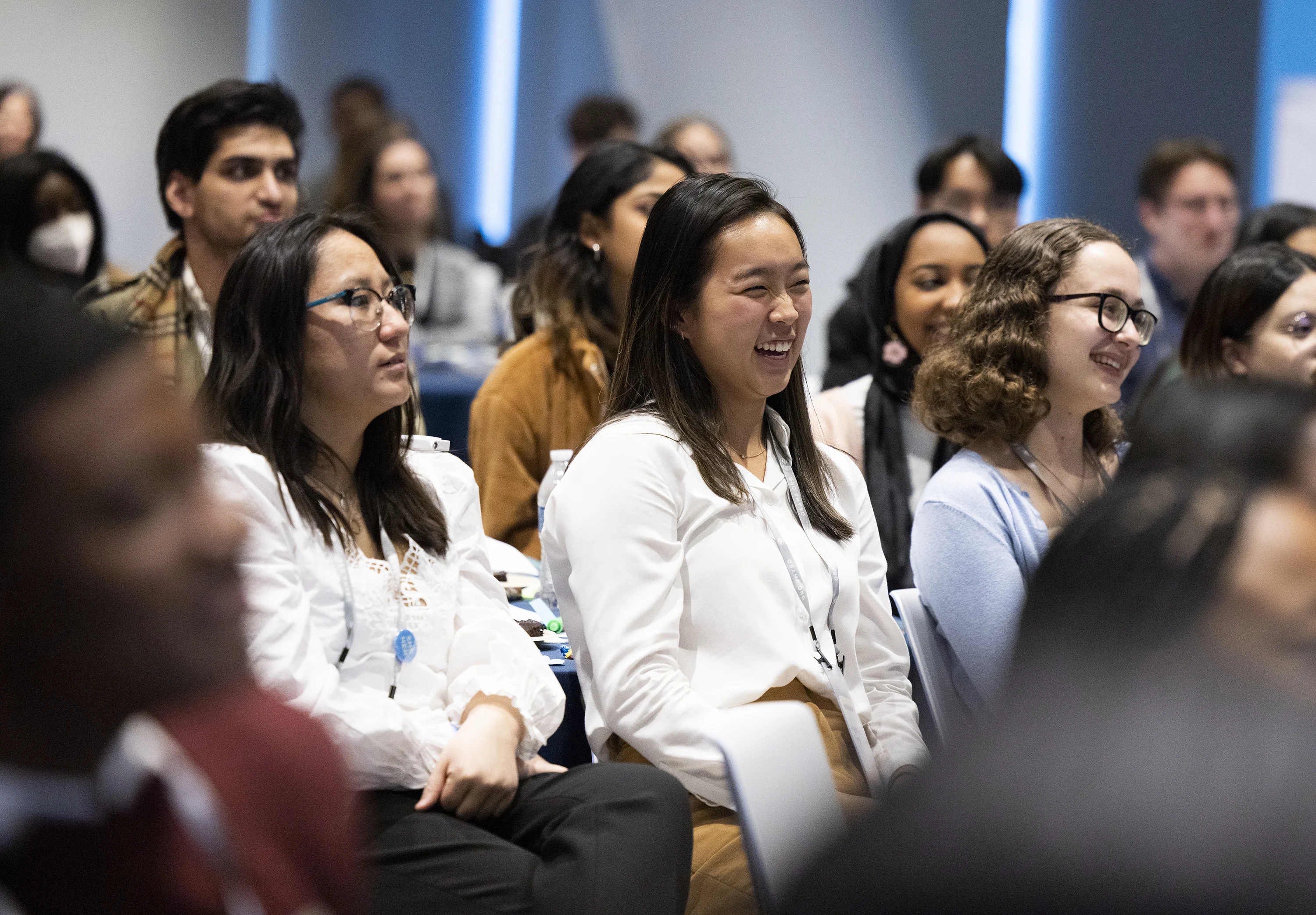 Alt text: A group of young people with a range of light to dark skin tones sit in a conference room. They are smiling and listening attentively. 