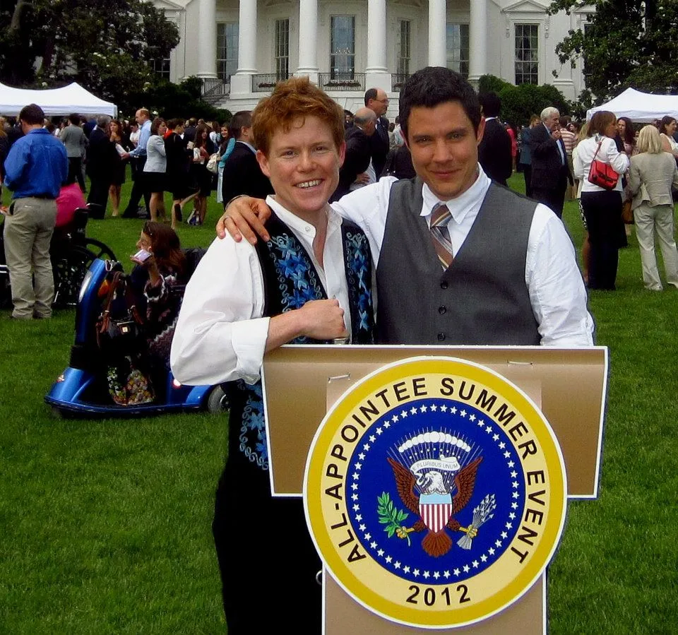 An image of Dylan Orr and a friend at a White House All-Appointee Summer Event in 2012. The picture is taken in the daytime on the White House Lawn. Dylan Orr and his friend stand close together behind a podium on the White House Lawn. Behind them are people sitting on green grass and the White House. Dylan Orr has an olive complexion with dark wavy hair. He is wearing a long sleeved white shirt, a gray vest, and a tie. His friend has pale skin and red shirt wavy hair. He is wearing a long sleeved white shirt and a blue vest. The podium they are standing behind is gold and has a large circular White House emblem on the front. On the emblem is the official presidential seal and the words, "All-Appointee Summer Event 2012"