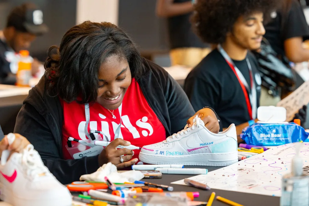 A woman with deep skin, long hair, and a nice smile designs an of Air Force 1s. She has a thin black jacket on and a red shirt that says "girls" on it. At the table she is working at are a bunch of craft supplies. On her shoe reads "I am powerful, Mariah." Other people are doing similar crafts out of focus in the background.