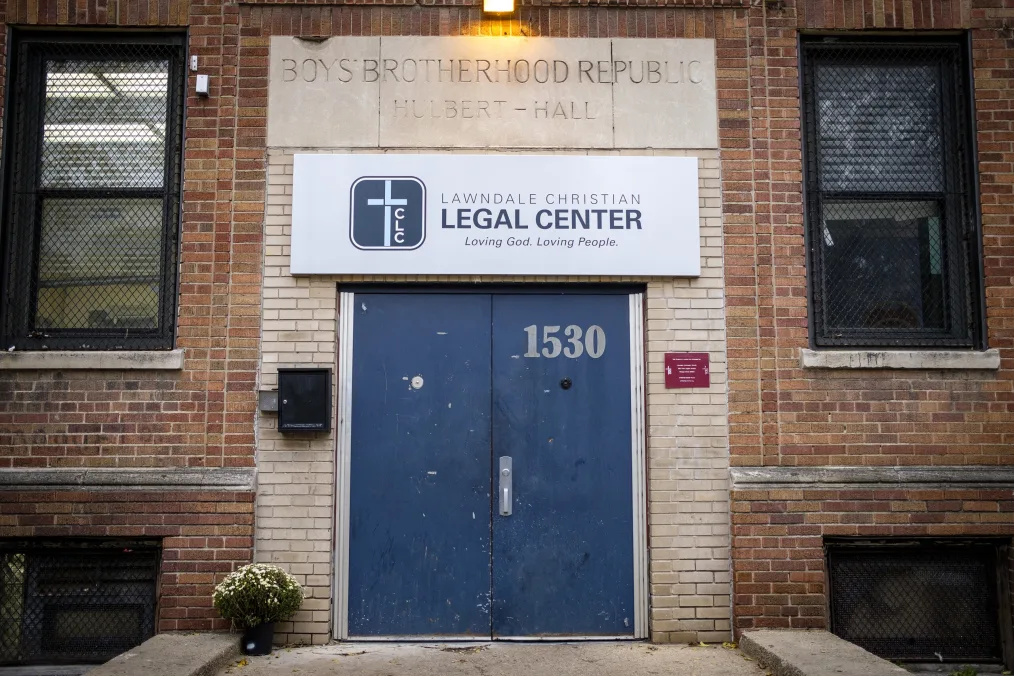 A photo of the entrance of a brick building. There are first floor and basement windows with black grated barriers on both sides of the door. The entrance has two blue steel doors and engraved in the stone above the doors reads "Boys Brotherhood Republic, Hubert-Hall." Underneath, on the supporting door frame is a board sign that reads "Lawndale Christian Legal Center, Loving-God. Lovinig People."