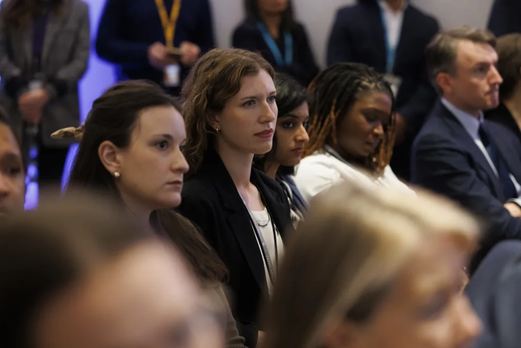 A group of individuas of a variety of skin tones wearing professional attire sit and stand in a room with their attention to the speaker or presentation. 