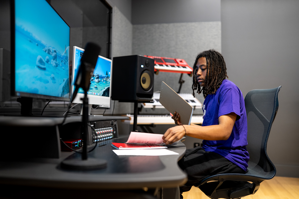 In a dark audio studio, a Black youth with dark dreadlocks sits in a chair at a curved desk. They look down at a folder and notebook. Large computer monitors sit on the desk on your left, along with a microphone and a speaker. Audio equipment sits in the background.
