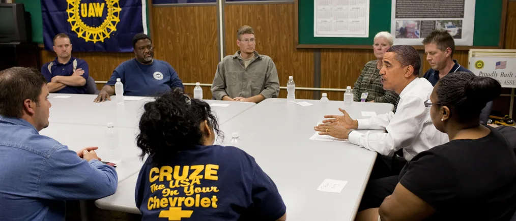 President Obama speaks to a table of people with various skin tones. The background features a blue flag with yellow lettering that reads ¨UAW¨
