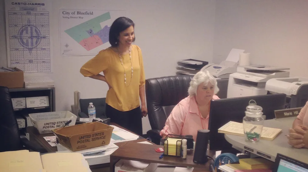 Two women look at a computer screen in an office with printers and copiers. One woman is sitting and one is standing behind her. There are bins with the words "United States Postal Service" on a desk in front of them and a map with the words "City of Bluefield Voting District" hangs on a wall behind them.