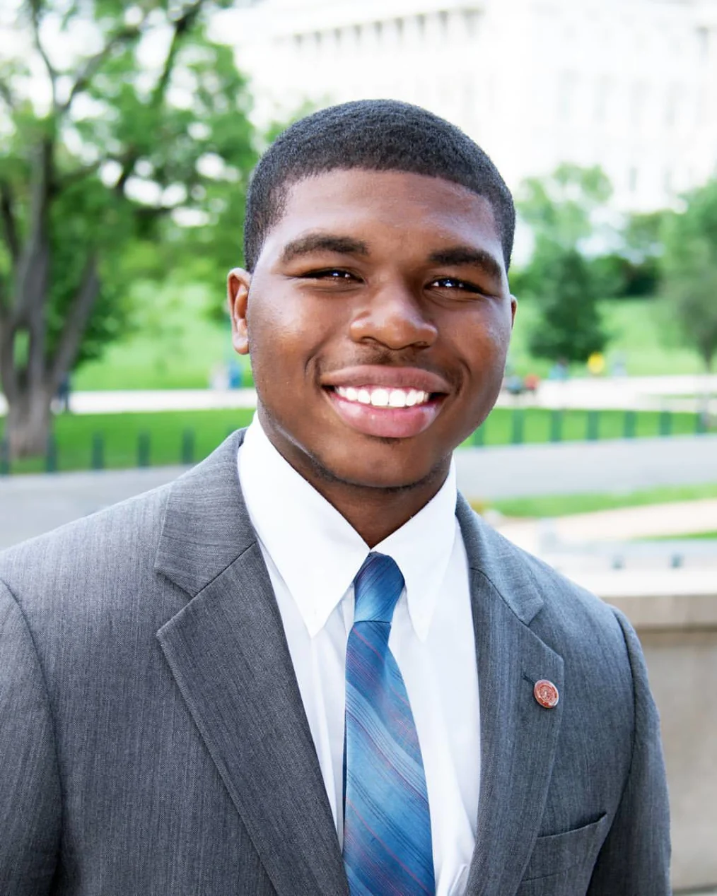 A chest up photo of a young man with medium-deep tone skin, low black hair. He has a nice white smile. He is wearing grey suit, with a white button up shirt underneath, and a tie thats pattern is a bunch of thin lines of blue shades. He is in focus of the camera while the background is a blue of a monument or museum park.  