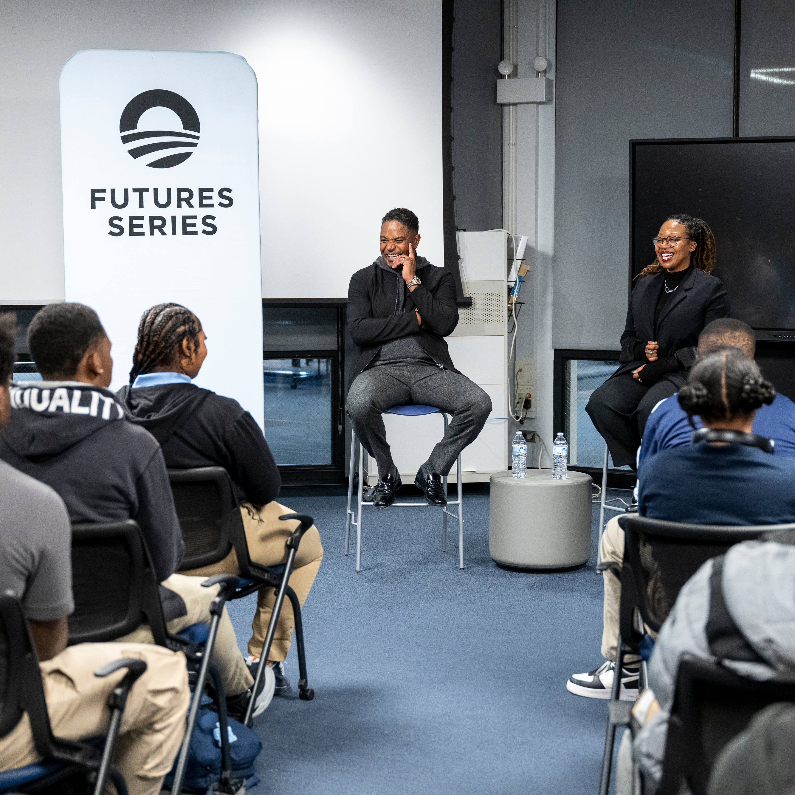 A man and a woman with medium skin tones are seated in front of a room of students with medium to dark skin tones. Their backs are to the camera. Two panels flank the speakers, one that says, “Futures Series” and one that says, “If you can see it, you can be it”