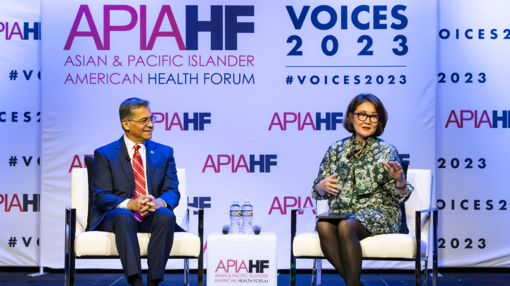The image is a candid photo of a panel at the 2023 Asian and Pacific Islander American Health Forum Voces 2023 Conference. In the photo two people are sitting on a stage in white chairs. Behind them is a white backdrop that has the logo for the Asian and Pacific Islander American Health Forum Voces 2023 Conference. The colors of the logo are pink and purple. In the first chair is a man. He has black and gray short hair and has an olive complexion. He is wearing glasses, a dark suit, a white button down shirt, and a red striped tie. He is smiling. The person in the second chair is Juliet Choi. She has dark black hair that is cut in a bob that hits the nape of her neck. She is of a lighter olive complexion and is wearing black rimmed glasses, a green blazer and dress with a white floral design on it and black stockings. She is talking and directing her gaze at the audience (the audience is unseen.) Between the two chairs is a white table. On the table are two deer park water bottles. On the front of the table is the Asian and Pacific Islander American Health Forum logo. 