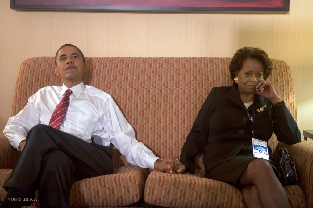 Barack Obama sits on a red and tan couch holding the hand of a woman (his mother-in-law Marian Robinson). They are both dressed formally, in suit attire. The two appear to be watching something intently. 