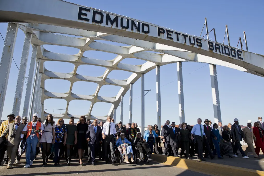 
President Obama and Michelle Obama walk under a bridge that reads " Edmund Pettus Bridge" with a diverse group of people ranging from young to old and light to deep skin tones 