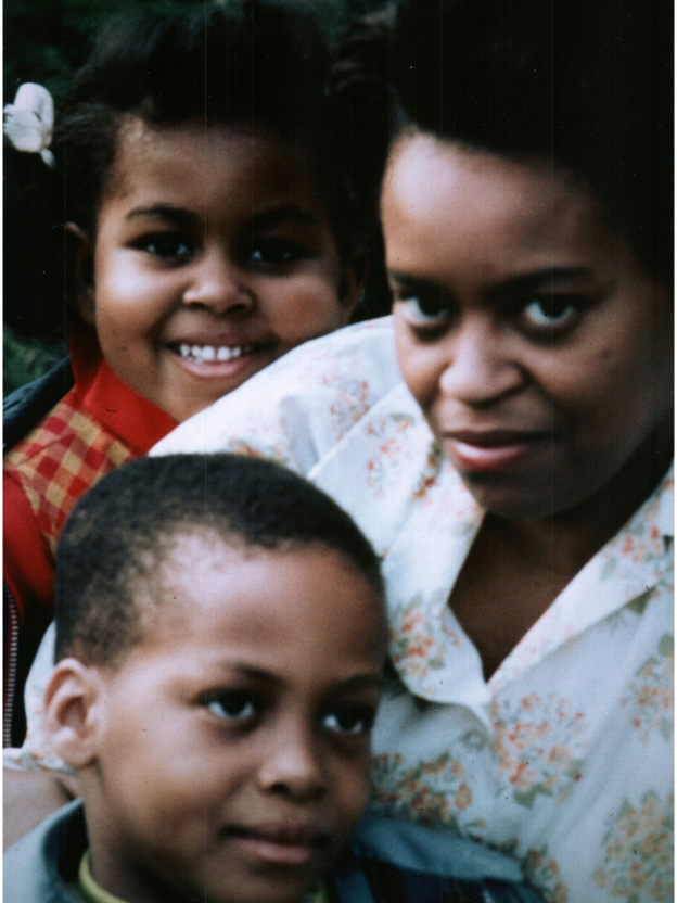 An image of Michelle Obama, Marian Robinson, and Craig Robinson from the 1970s. Michelle Obama is a child: she has a bow in her hair. Marian Robinson is younger: she is wearing a white shirt with flowers on it. Craig Robinson is a young boy: he is smiling and looking away from the camera.
