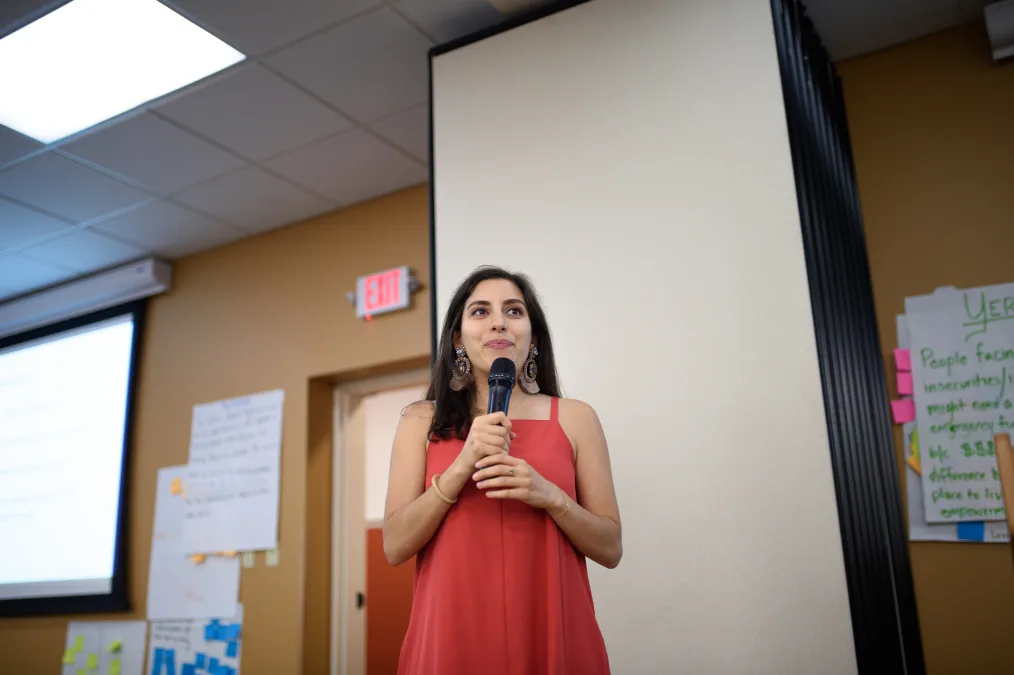 A woman with a light-medium skin tone, a red dress, and big brown designed earrings stands in front of a golden-tan wall, filled with paper with blue, pink, and yellow sticky notes and writing, holding a microphone in her hand up to her mouth. 