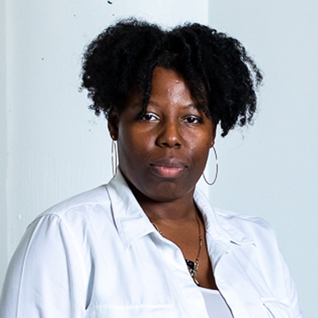 Tyanna is sitting with her legs crossed on a dark colored chair staring into the camera. Behind her is a white wall with a green tapestry hanging from one side. Next to the wall is a small stack of bricks. Tyanna has a deep skin complexion and medium length black hair. She is wearing hoop earrings, a white long sleeved shirt and a white skirt. On her feet are brown loafers.