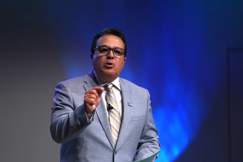 Dr. Edwin Quezada, a man with a medium skin tone points as he speaks to an audience. He is wearing glasses and a powder blue suit. 