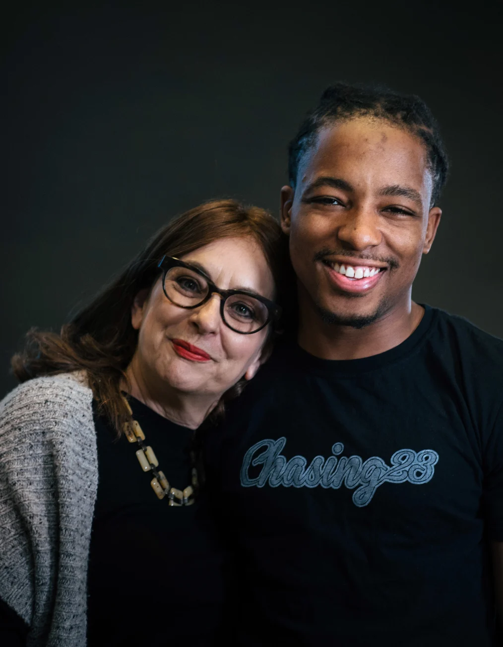 This portrait shows a man with a medium-deep skin tone wearing a black shirt and smiling toward the 
camera. Next to him is a lady with a light skin tone wearing a gray sweater, glasses, and a necklace, 
smiling toward the camera.