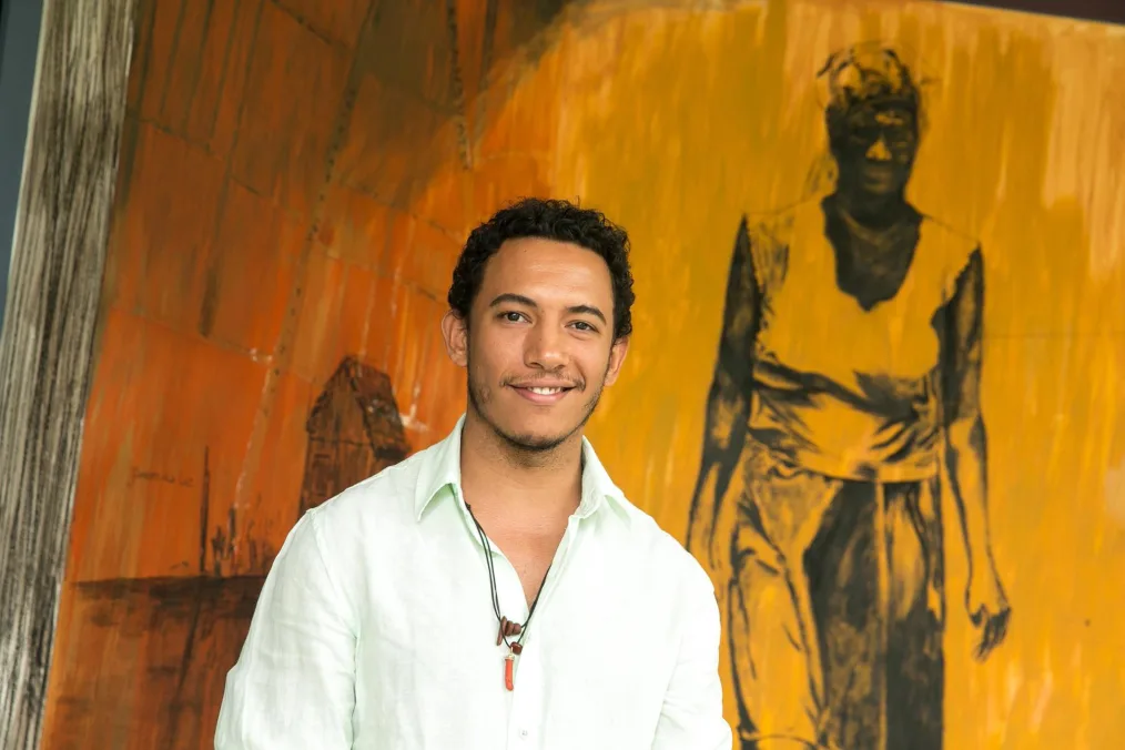 A person with medium skin tone and curly dark hair smiles at the camera. He wears a light green button-up shirt with the first few buttons undone and two black rope necklaces. He stands in front of what appears to be a painting in reds, oranges and yellows, of a person wearing loose clothing walking.