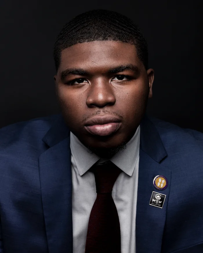 A portrait photo of a young man with a deep skin tone wearing a white button-up shirt, a burgundy tie, and a navy blue blazer with white "MBK Alliance" stitching and a round yellow, white, and purple pin on the collar standing in front of a gradient black background. 