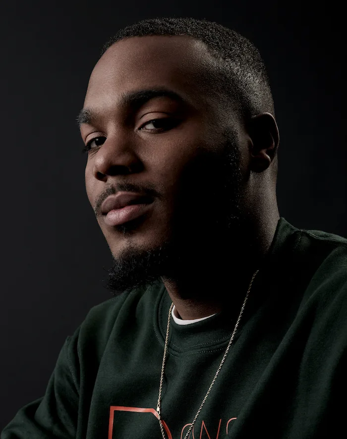 A portrait photo of a man with a deep skin tone wearing a dark green sweatshirt in front of a gradient black background. 