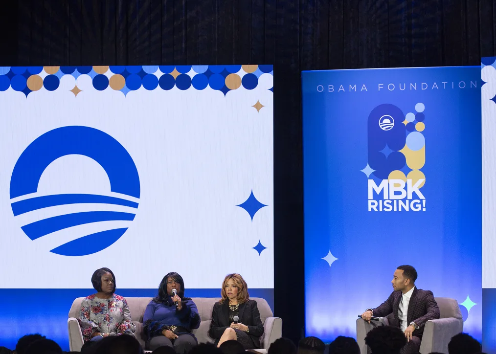 John Ledgend speaks with a panel of black women with various skin tones speak to an audience. The background feature orange, white, and blue ¨MBK Rising!¨ and Obama Foundation graphics.