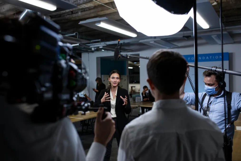 Nima Tisdall stands in front of a man holding a video camera, and a man holding a sound boom, gesturing with her hands as she looks forward.