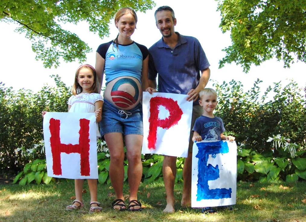 A pregnant woman wearing an "Obama '08" t-shirt and with the Obama Rising Sun "O" logo painted on her belly, a man in a collared shirt and two young children hold up painted signs that spell the word "Hope." Behind them are trees and shrubs.