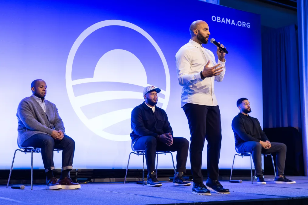 Jeremy Watson, a Black man with a light skin tone and no hair, stands center stage and speaks to a crowd. Behind him, sitting, are three other Black men with a range of light to medium skin tones. An Obama Foundation rising sun logo is in the background.