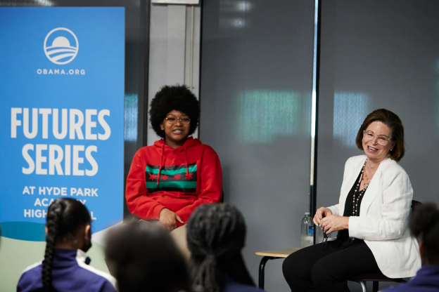 Valerie Jarrett sits smiling and speaking to a crowd of young students with dark hair. Sitting beside her, a young person with a medium deep skin tone who wears a red sweatshirt that displays a red, green and black Chicago flag. Next to them, a banner that reads 'FUTURES SERIES AT HYDE PARK ACADEMY HIGH SCHOOL." 