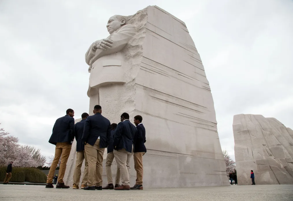 Young men gather in front of the Martin Luther King, Jr. Memorial. All have a range of light to deep skin tones and are wearing suit jackets and khaki pants.