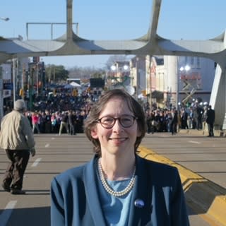 A candid shot of Pamela Karlan. Pamela is standing outside in front of a large crowd of people. Pamela has pale skin and brown hair cut to her shoulders. She is wearing round glasses, a pearl necklace, and a dark blue blazer and shirt. She is smiling into the camera.