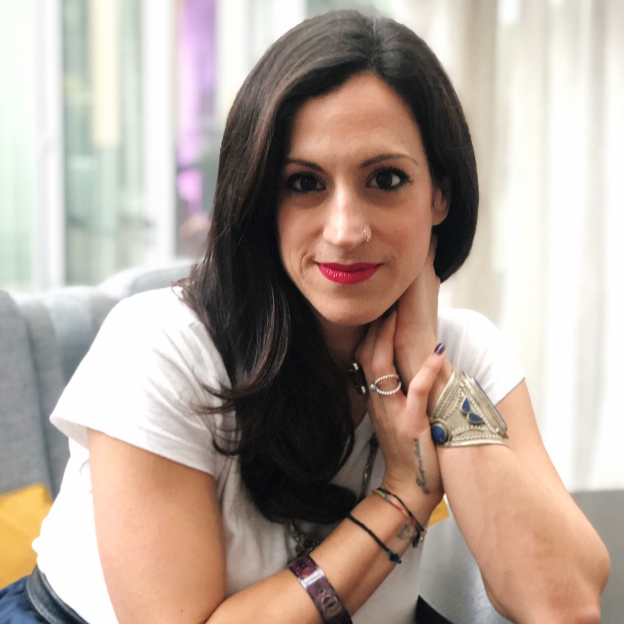 Elpida Kokkota, a woman with a light skin tone, holds a closed lip smile. She has long black hair and is wearing red lipstick, bracelets on both arms, a white shirt, and floral skirt. 