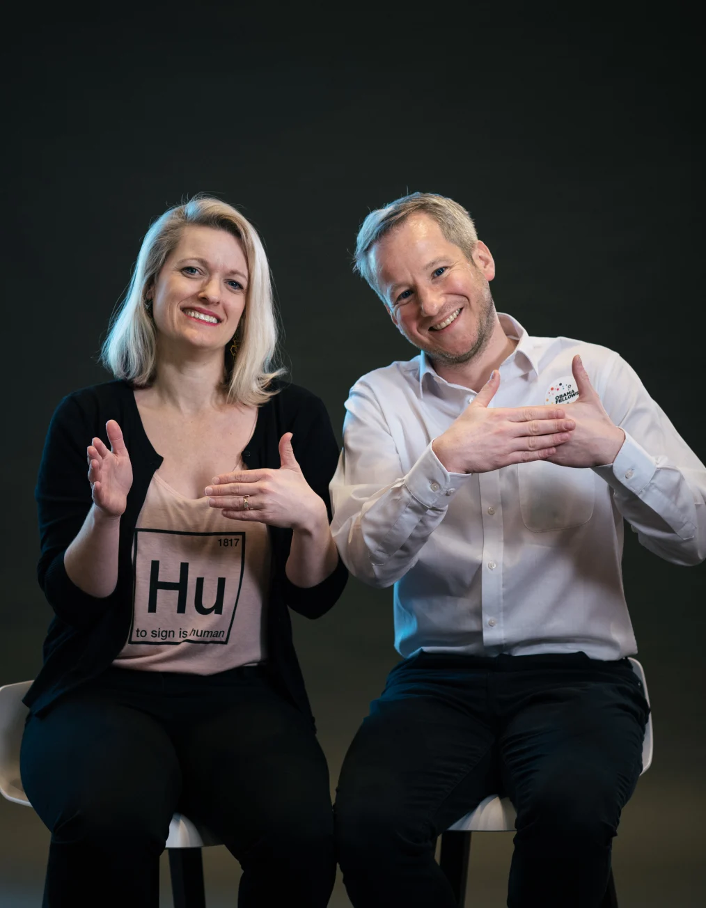 In this picture, a man and woman with a light skin tone are shown sitting down on white chairs 
smiling and gesturing a hand sign toward the camera in front of a black backsplash.