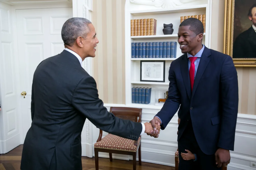 The image is a candid shot of Jeron Smith shaking the hand of President Barack Obama. The location of the shot is the White House Oval Office. On the wall of the office is a beige striped wallpaper and a white bookshelf holding gold and blue books. On the bookshelf are additional presidential artifacts. To the right of the bookshelf is a portrait of Abraham Lincoln to the left of the bookshelf is the door to the Oval Office. Underneath the bookshelf is a brown wooden chair. In the center of the image is Jeron Smith and President Barack Obama shaking hands. On the left is President Obama: he has light brown skin, closely cut black and grey hair, and is wearing a dark gray suit with a white shirt underneath. He has his right arm outstretched to towards Jeron. On the right side is Jeron Sminth: he has brown skin, closely cut dark hair, and is wearing a dark blue suit with a light blue shirt and red tie underneath. He has his right arm outstretched to President Obama and is smiling. At the very bottom of the picture is a small child holding onto the back of Jeron's right leg. The child is hiding behind Jeron and is not visible to the camera.