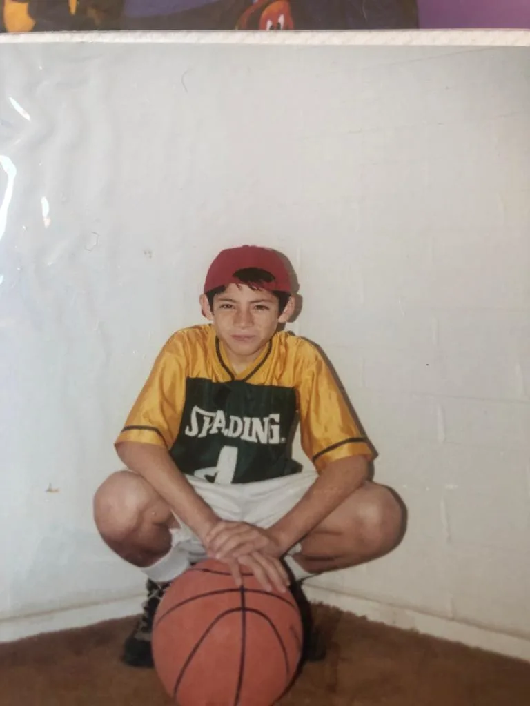a young José Patiño squats down in front of a basketball. He is wearing a red hat and yellow Spaulding jersey.