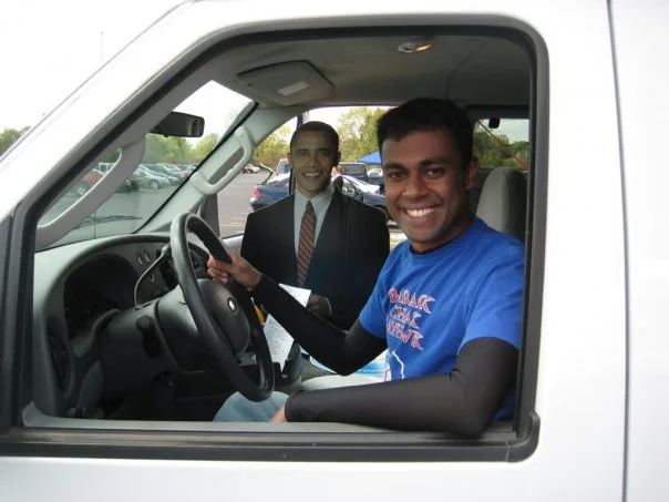 Vineeth Hemavathi, sits in the driver’s seat of a white pick-up truck smiling at the camera. He is wearing a blue t-shirt with red lettering that says “Barack Chalk Jayhawk.”' Next to him in the passenger seat of the white pick-up truck is a full scale cardboard cutout of Barack Obama. The image is of Barack Obama when he was an Illinois Senator. In the background of the pickup truck is an outdoor parking lot.  