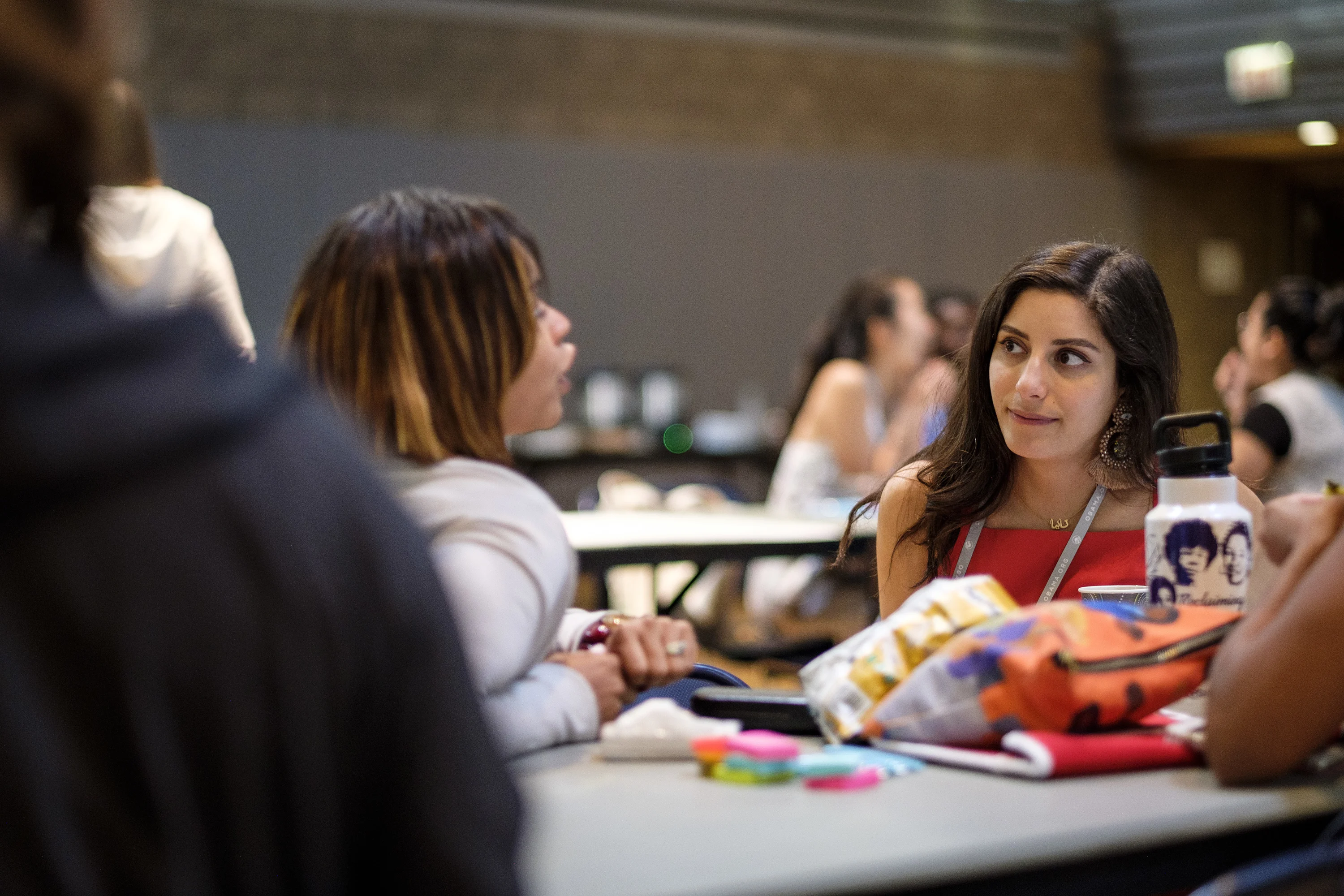 A woman with a light-medium skin tone and a red shirt is sitting at a table next to another woman with a light-medium skin tone and a light gray sweater. The woman with the light gray sweater is talking. There are pouches, small supplies, and a water bottle on the table. There are other people blurred in the background. 