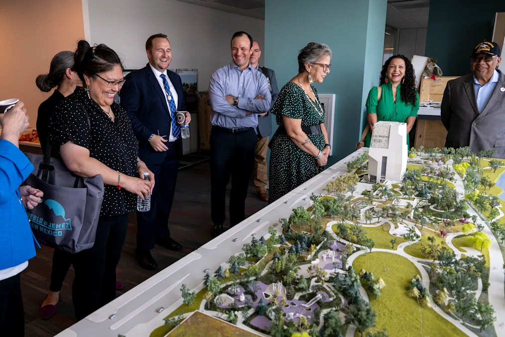 A photo of Charlie Galbraith standing with several Chicago Indigenous leaders in the Obama Foundation Office located in Chicago, Illinois. Charlie and the leaders are looking at a small-scale replica  of the Obama Presidential Center. The small-scale replica features the exterior of the space and has fake grass and small icons representing each building on the campus. In the image are eight people including Charlie. Charlie is standing in the center of the group. The people have a variety of skin tones and are all wearing a variety of outfits. They are all smiling in the picture. 