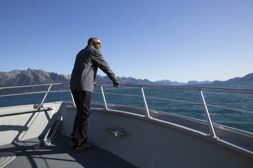 A side view of President Obama wearing black shades, a black jacket, and black pants, He at the bow of a boat surrounded by water around.