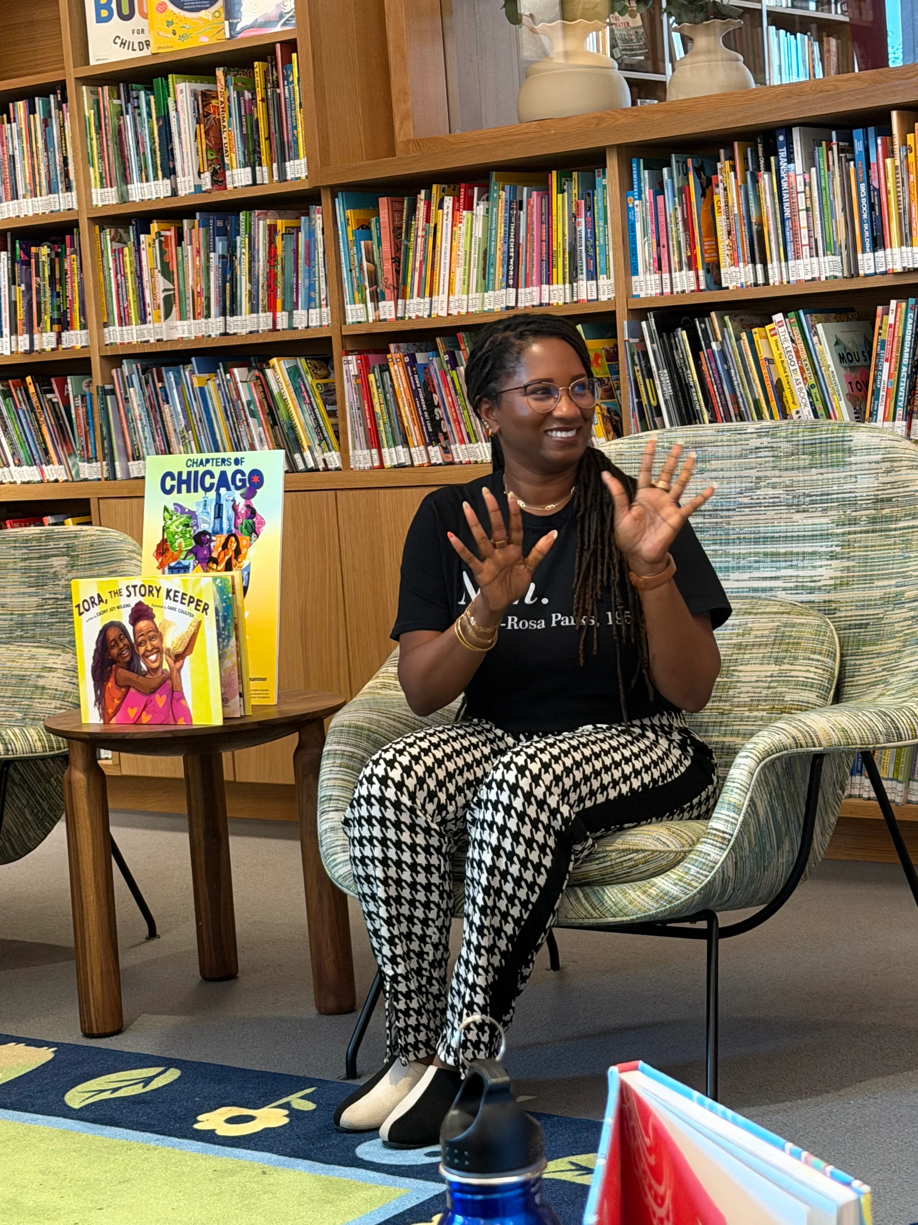 Ebony Wilkins, A Black woman with a dark skin tone and box braids, sits as she reads to a group of children with a range of light to dark skin tones at the Chicago Public Library Branch at the Obama Presidential Center. Her hands are spread out. She is smiling.

