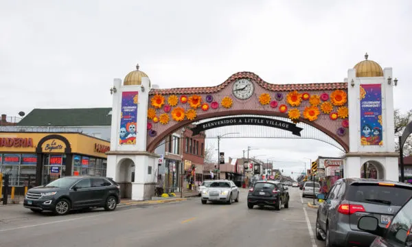 A colorful archway with a clock in the middle and orange and pink flowers spans a road with cars driving down it. The words "Bienvenidos a Little Village" are on a sign beneath the archway.