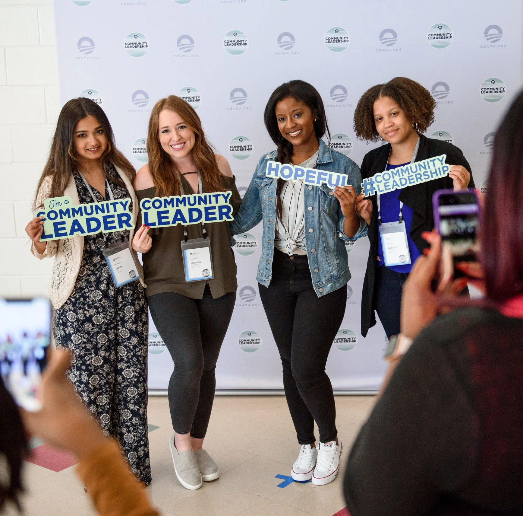 Four women with a variety of different skin tones stand in front of a light blue Obama Foundation backdrop holding community and Obama Foundation signs. There are blurred people taking pictures of them.