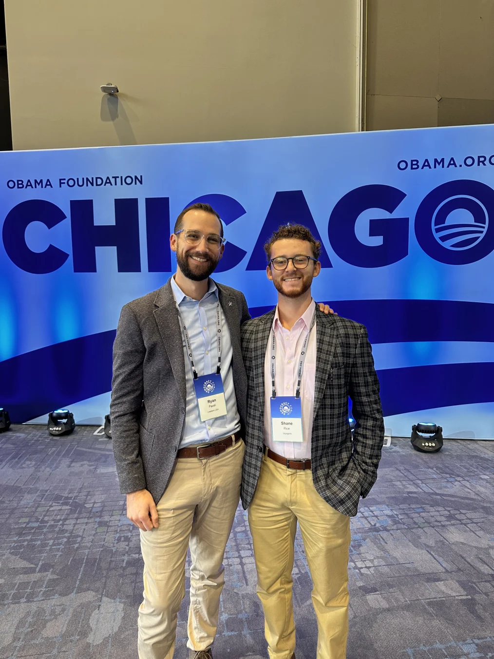 Standing in front of a large blue photo backdrop that reads, “Chicago” in all caps, Ryan Patel and Shane Rice, two white males dressed in business casual attire, have their arms around each other for a photo. Both are wearing blazers. 