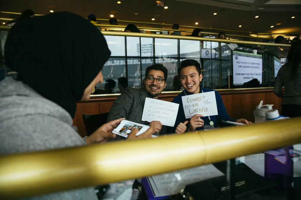 In this picture, two men and a female with medium skin tones are shown inside a room at a table. The
female is taking a picture of the two men posing toward the camera holding signs.