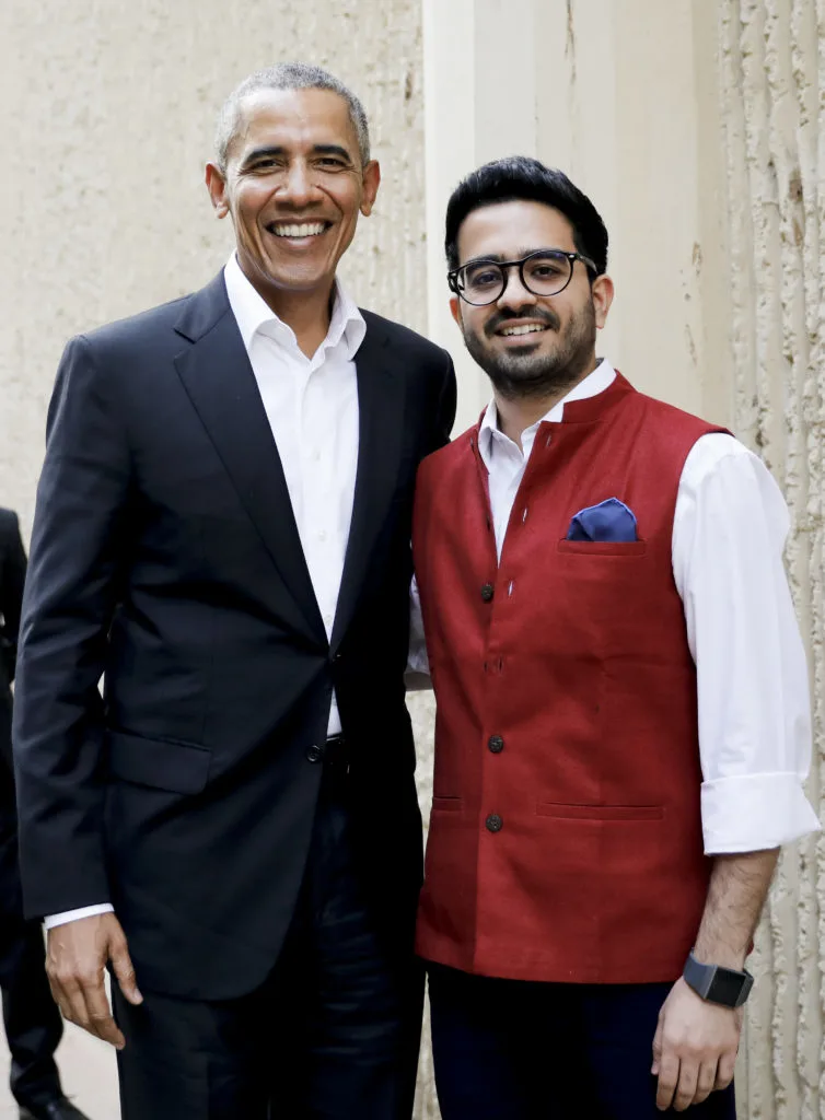 President Obama stands side-by-side with a man who has a light medium skin tone with combed, slicked back hair, glasses, scruffy beard and moustache, and a white button up shirt with a red vest.