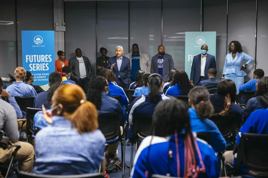 A group of students in matching blue jackest partake in a furture series meeting hosted by the Obama Foundation. A line of darker toned presenters in formal attire stand behind a light-medium toned man in formal attire leading the talk.  
